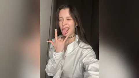 Playful young woman in a white button-down shirt making a hand gesture and sticking her tongue out indoors.