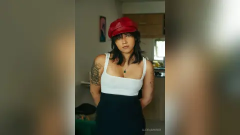 Young woman in a white tank top and red beret posing in a modern kitchen with a confident mood.