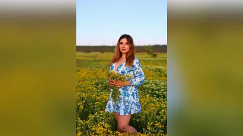 Young woman in a blue floral dress standing in a sunny yellow wildflower field under a clear blue sky.