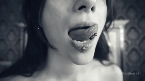 Close-up portrait of a woman with tongue and chin piercings in a moody black and white setting.