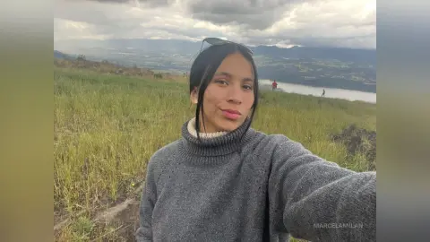 Young woman in a grey turtleneck sweater smiling in a grassy field with mountains under a cloudy sky.