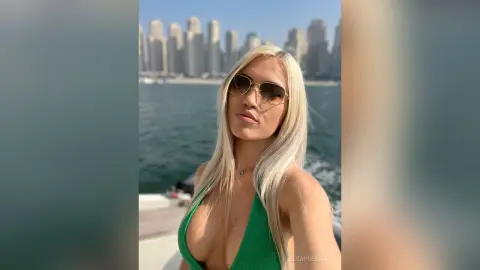 Blonde woman in a green bikini sunglasses enjoying a sunny summer day on a boat near a city skyline.