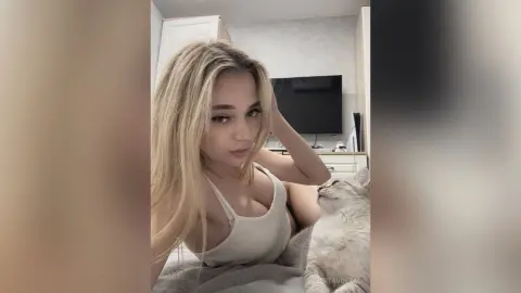 Blonde woman in a white tank top relaxing at home with her grey cat in a cozy, bright bedroom setting.