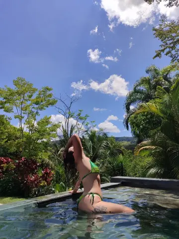 Woman in green bikini relaxing in a tropical outdoor swimming pool under a sunny blue sky.