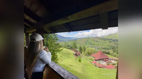 Woman in a white jacket looking out from a wooden balcony at a scenic green mountain valley landscape.