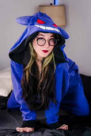 SallyLynn18 wears a blue Gengar onesie and glasses while posing on a bed with her long dark hair.