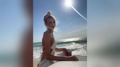 Blonde woman in a white crochet bikini relaxing on a sunny beach under a bright blue sky.