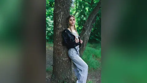 Blonde woman in a black leather jacket and light denim jeans leaning against a tree in a lush green forest.