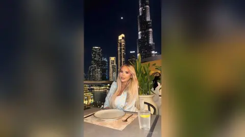 Blonde woman in a white blouse dining at a rooftop restaurant overlooking the Dubai skyline at night.