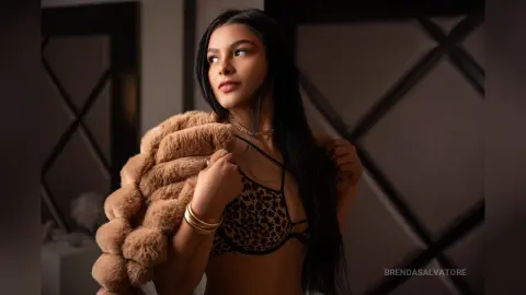 Young woman in leopard print bikini and brown faux fur coat posing in a moody, dimly lit indoor setting.