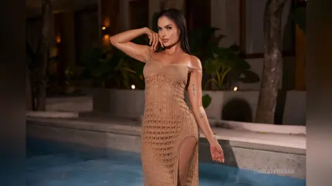 Elegant woman in a beige lace slit maxi dress posing by a luxury swimming pool at night.