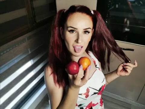 Playful woman with red pigtails in a floral dress holding peaches in a bright indoor setting.