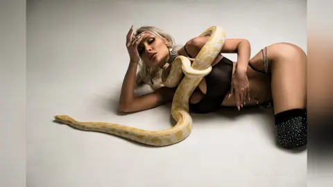 LeslyAnderson lies on a white floor wearing a black bodysuit, posing with a large light-colored snake draped over her body.