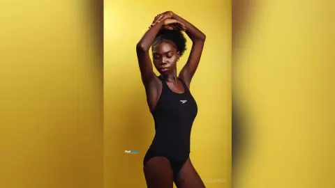 Confident Black woman in a black Speedo swimsuit posing against a vibrant yellow studio background.