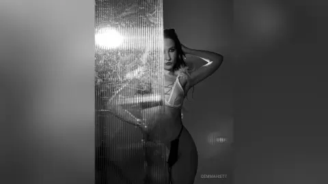 Moody black and white portrait of a woman in white lingerie posing behind a textured glass partition.
