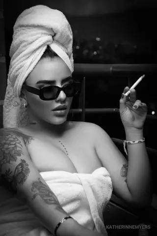Tattooed woman in a white towel and sunglasses smoking a cigarette on a balcony at night.