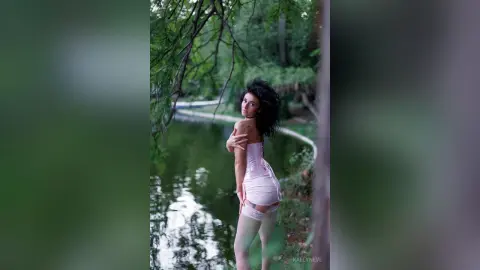 Dreamy portrait of a woman in a pink satin slip dress standing by a serene forest pond.