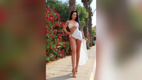 Elegant woman in a white two-piece outfit posing on a sunny garden path by red flowers.
