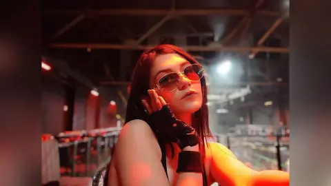 Young woman in black sleeveless top and sunglasses posing in a moody, dimly lit urban setting.