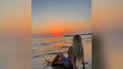 Blonde woman in a colorful floral dress sitting on a beach at sunset watching the ocean waves.