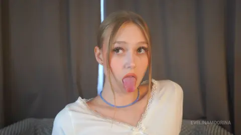 Blonde woman in a white lace top sticking her tongue out playfully in a cozy indoor setting.