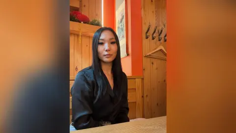 Young woman in a black blouse sitting in a warm, rustic wooden room with a calm expression.