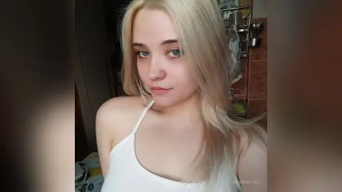 Blonde woman in a white tank top posing with a soft, natural look in a cozy indoor setting.