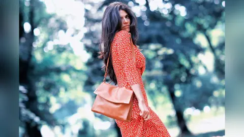 Woman in a red polka dot summer dress with a tan leather crossbody bag walking through a lush green forest.