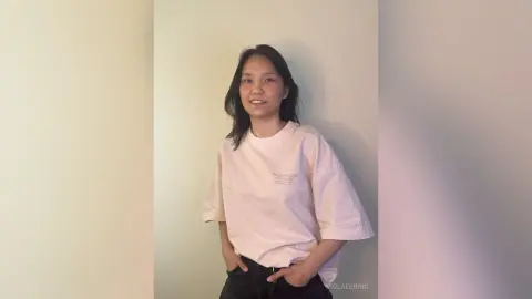 ViolaEbbing is a smiling woman with long dark hair wearing a loose white t-shirt and black pants, standing against a plain wall.