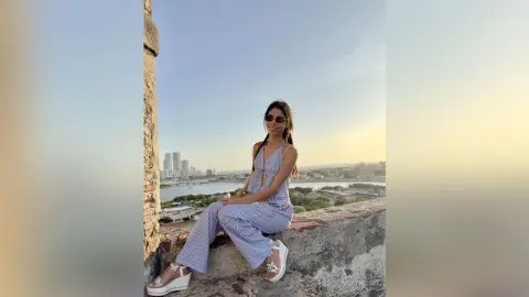 Smiling woman in blue striped jumpsuit and white sandals sitting on a rooftop overlooking a city skyline.
