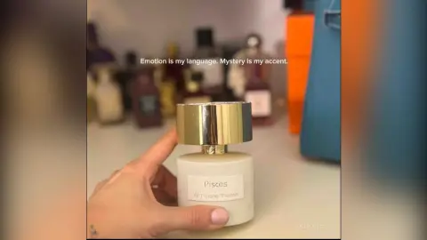 AnaMathers holds a white bottle of Pisces perfume against a blurred background of other glass bottles.