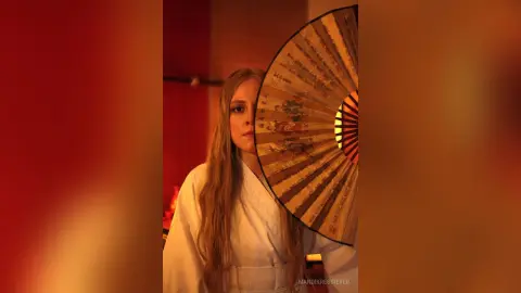 Blonde woman in white robe holding a traditional paper fan in a warm, moody red-lit setting.
