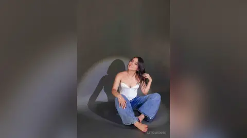 Young woman in a white corset top and blue jeans sitting on a studio floor with dramatic shadow lighting.