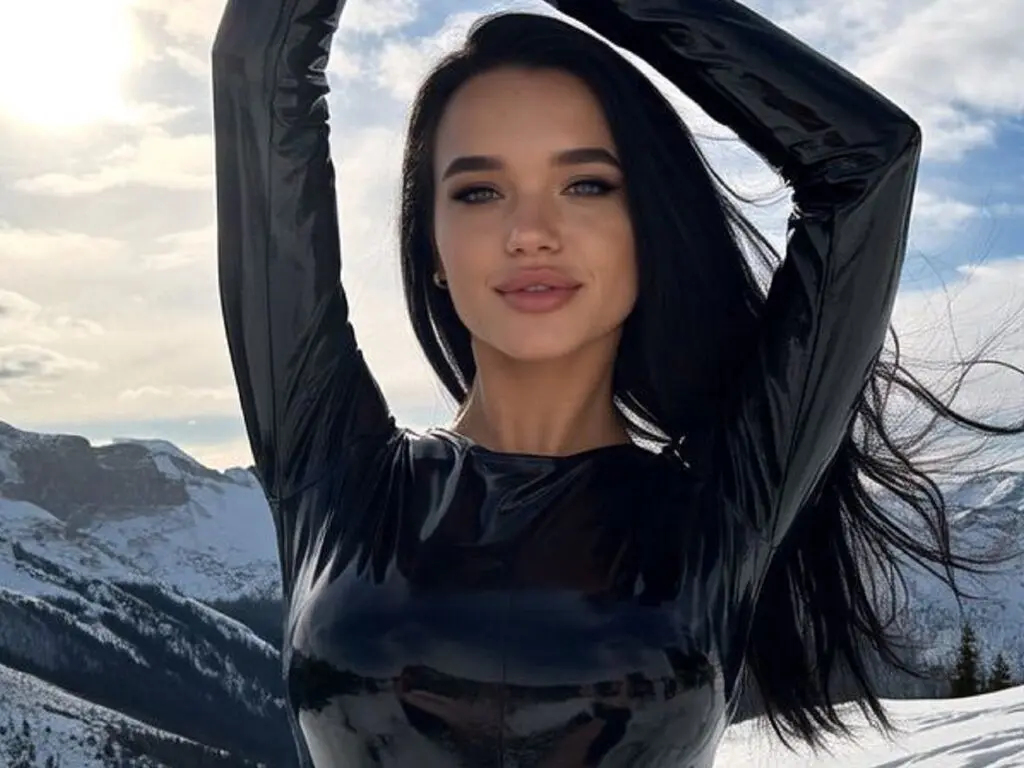 Young woman in a black shiny latex top posing against a snowy mountain landscape under a bright sky.