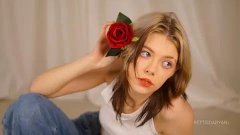 Young woman in a white tank top with a red rose behind her ear, posing in a soft, dreamy studio setting.