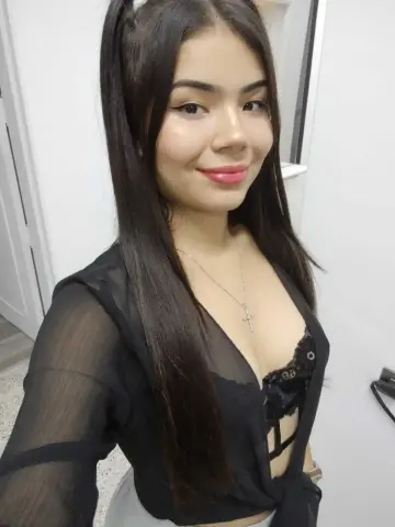 Smiling young woman with long dark hair wearing a black sheer blouse and silver cross necklace indoors.