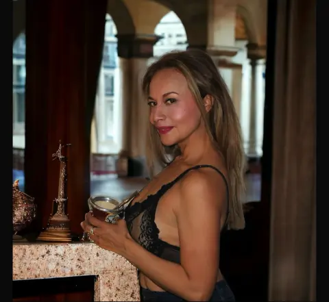 Smiling blonde woman in a black lace top holding a drink in a classic, moody architectural setting.
