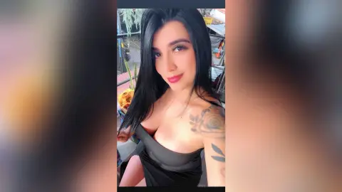 Smiling woman with long dark hair and floral tattoos wearing a black dress in a bright outdoor setting.