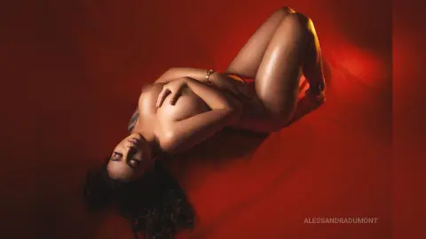 AlessandraDumont lies naked on a red sheet, covering her breasts with her hands and posing with her legs bent.