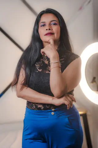 Elegant woman in a black lace top and blue trousers posing thoughtfully in a bright, modern room.