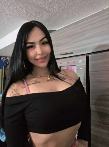 Smiling woman with long dark hair wearing a black off-the-shoulder crop top in a cozy indoor setting.