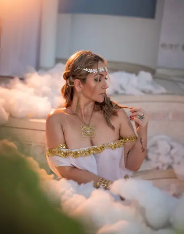 Ethereal woman in white goddess dress and gold butterfly necklace resting in soft white clouds.
