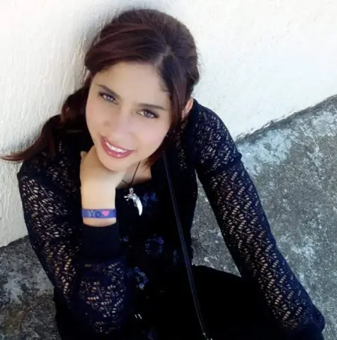 SilvannaReed smiles while leaning against a white wall, wearing a black lace long-sleeve top and a purple wristband.
