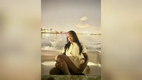 Elegant woman in a white knit top sitting on a boat at night overlooking a glowing city skyline.