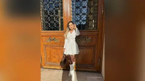 TiffanyAddams stands in front of a large wooden door wearing a white button-up dress and white boots, holding a brown handbag.
