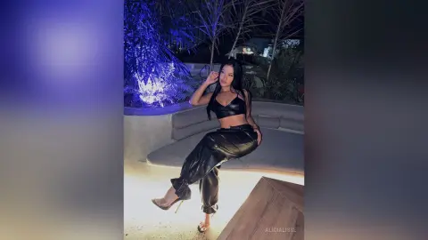 Young woman in black leather crop top and trousers sitting on a lounge bench in a moody outdoor night setting.