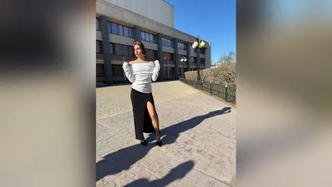 Elegant woman in a white off-the-shoulder top and black slit skirt standing on a sunny urban walkway.