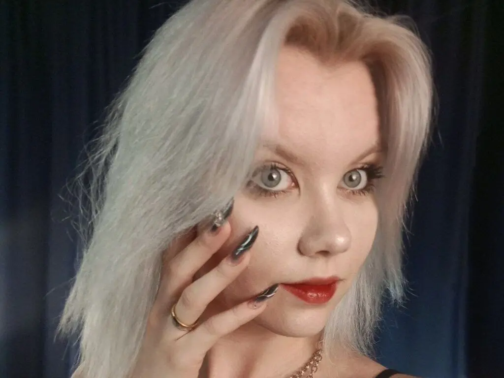 Close-up portrait of a fair-skinned woman with platinum blonde hair and dark nail polish against a blue backdrop.
