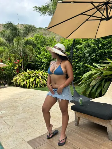 Woman in blue bikini top and denim shorts relaxing under a patio umbrella in a tropical garden setting.