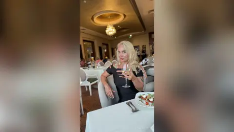 Blonde woman in a black dress enjoying white wine at an elegant restaurant dinner setting.